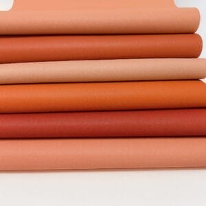 Folded hides of Milano cowhide leather in coral, orange, and blush tones, displayed on a white background. The article features a soft texture and subtle natural grain, ideal for leathercraft and accessories.-Avetco